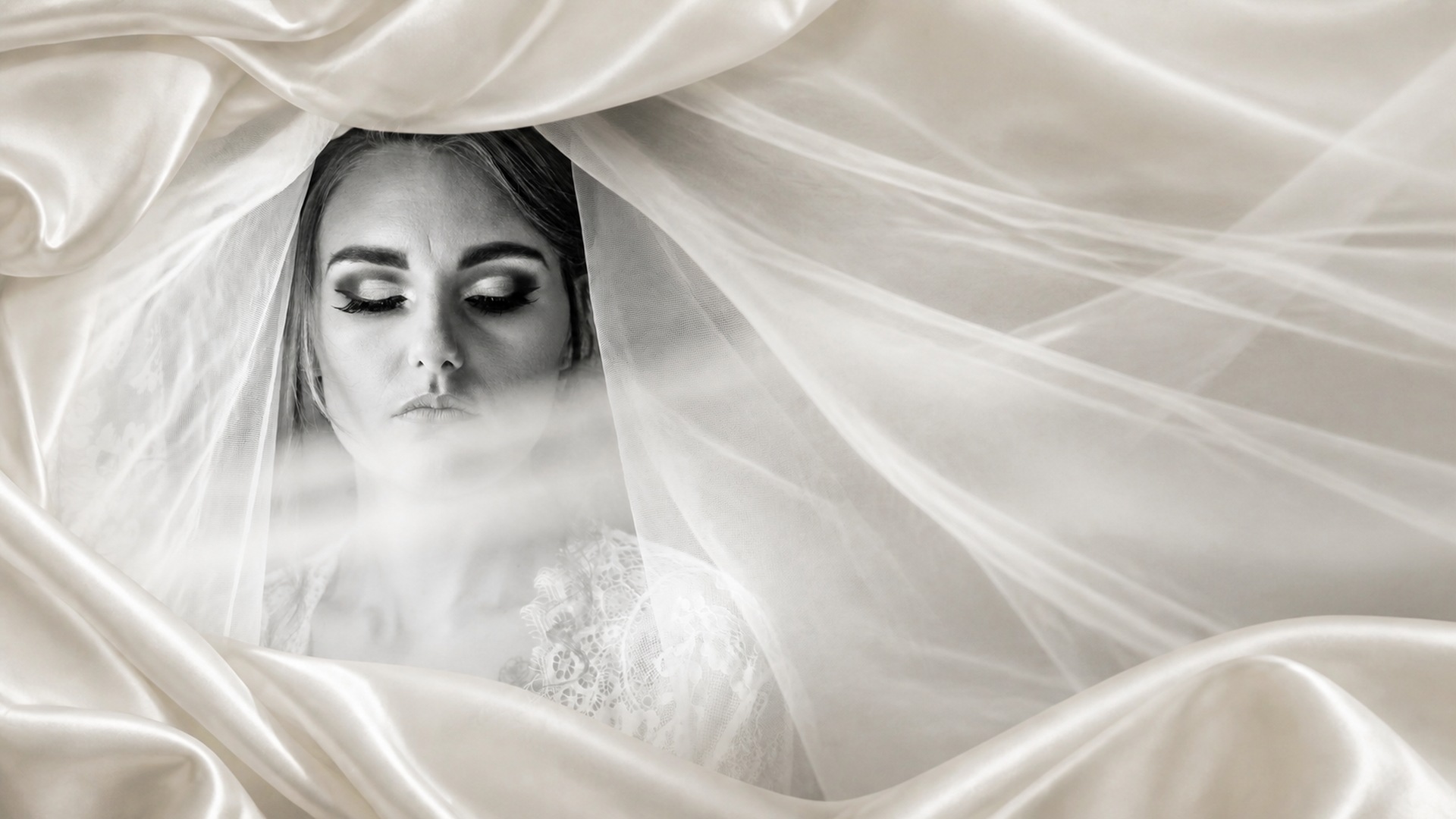 Black-and-white portrait of a bride surrounded by ivory satin and translucent veil